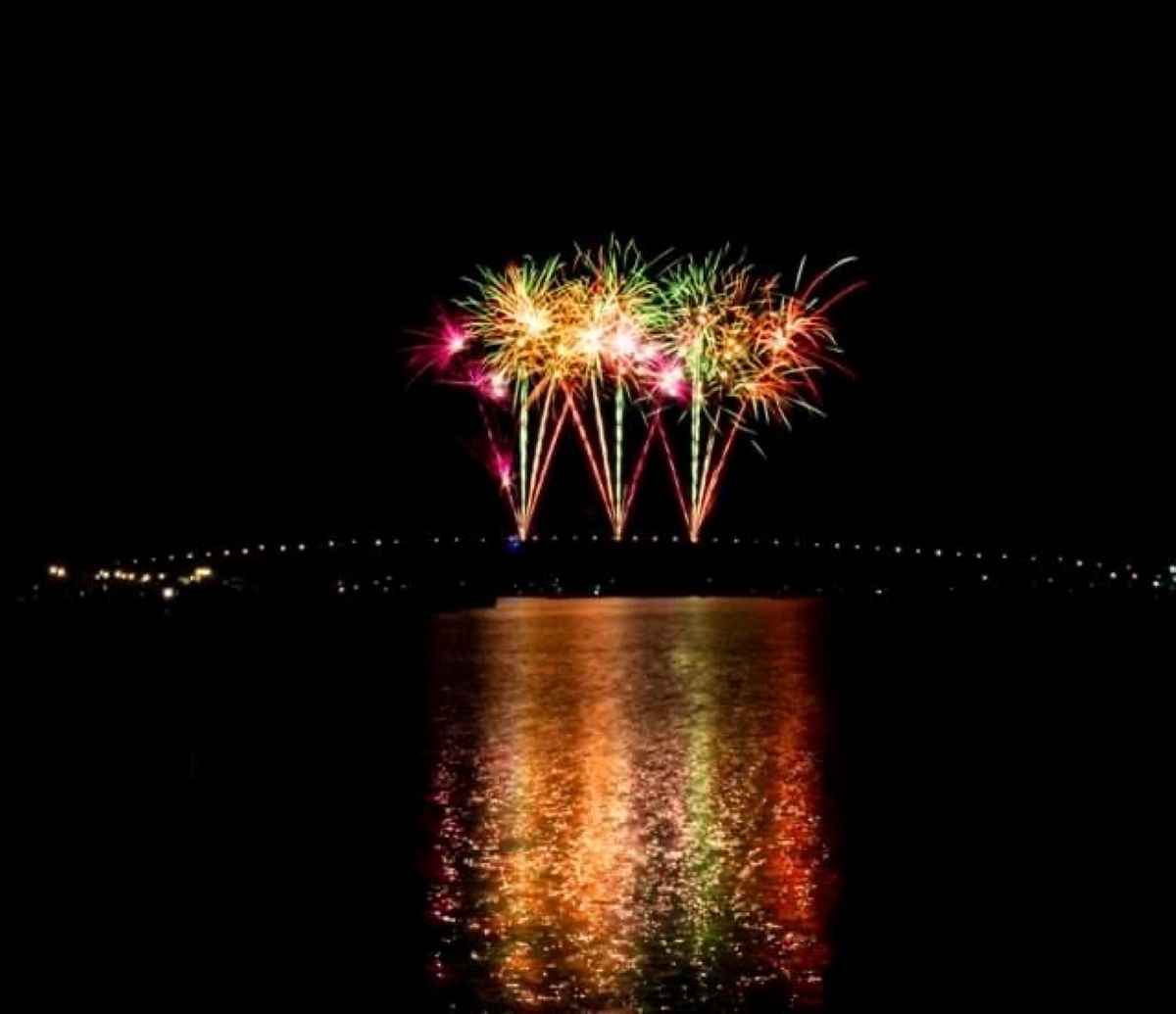 Colourful fireworks burst over a dark bridge, reflecting on the water below.