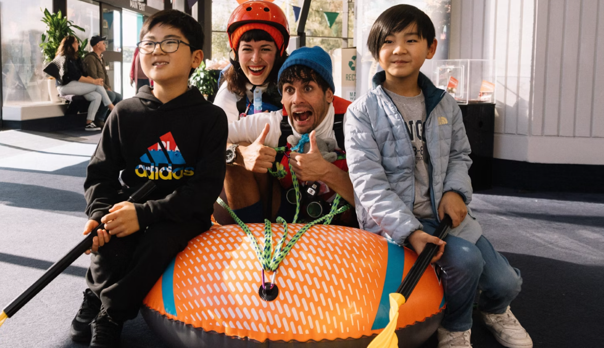 Two kids and two adults with helmets pose with a raft indoors, smiling and giving thumbs up.