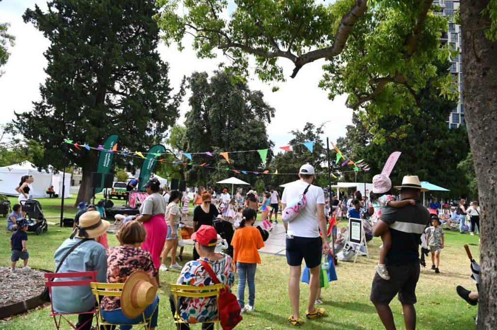 A diverse crowd of people seated on chairs, enjoying a sunny day in a vibrant park setting.