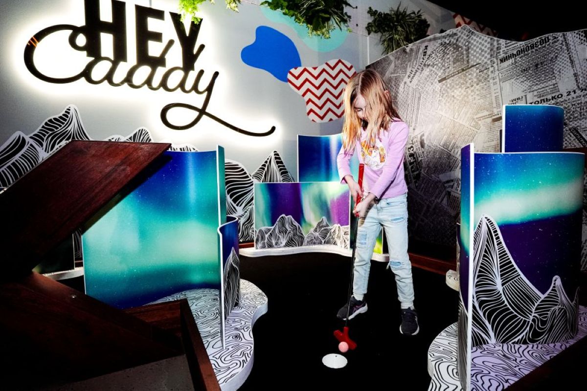 A young girl smiles while playing mini golf indoors, with a playful sign reading "Hey Caddy" in the background.