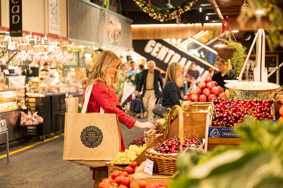 A woman shops for fresh fruits and vegetables at a bustling market, surrounded by colorful produce displays.