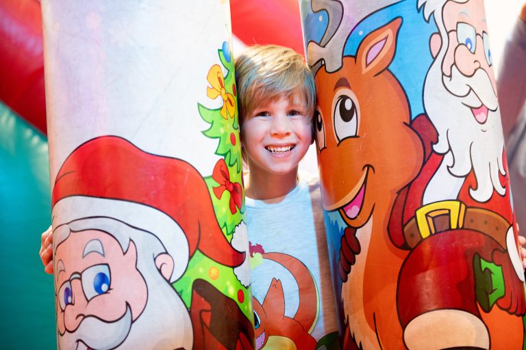 Boy smiles at camera, as he stands between inflatable Santa and reindeer.