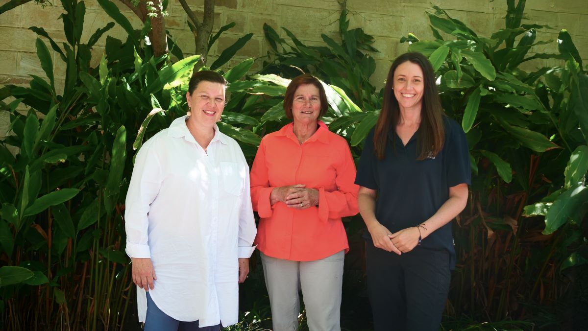 Three women stand in front of a plant.