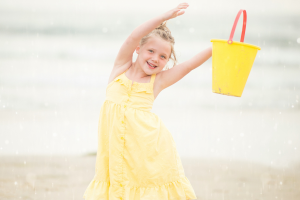 A little girl in a yellow dress joyfully holds a bucket, ready for a fun day of play and adventure.