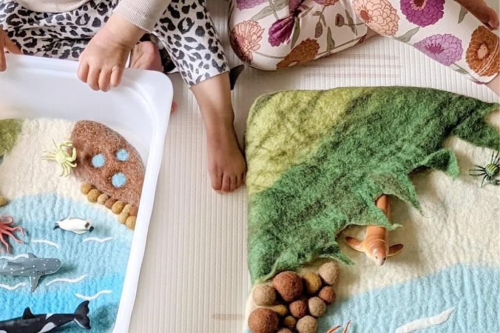 Two children joyfully playing on a colorful felt ocean-themed bath mat, surrounded by sea creatures and waves.