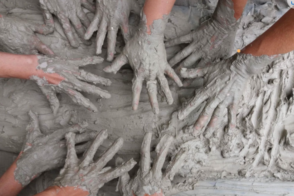 A group of hands from different people working together to mold a large pile of clay.