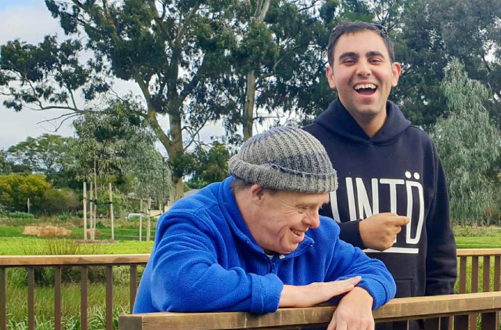 Two men sitting on a wooden fence, sharing a laugh and enjoying each other's company in a sunny outdoor setting.