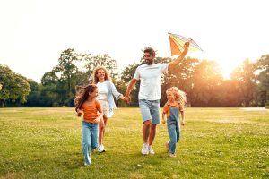 A family runs in the park, flying a colourful kite on a sunny day.