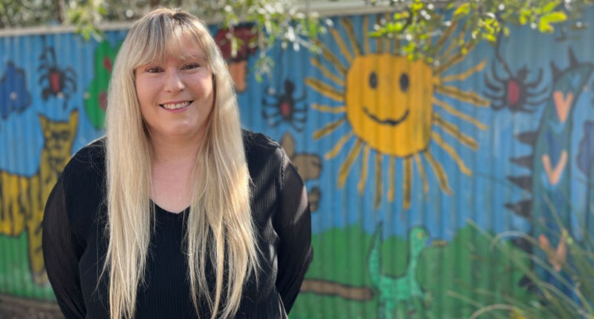 A woman with long blonde hair stands confidently in front of a vibrant, colorful wall.