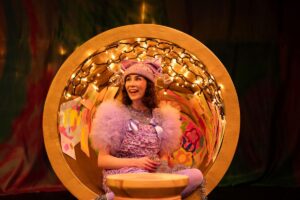 A woman in a purple costume sits gracefully in a chair, exuding elegance and charm.