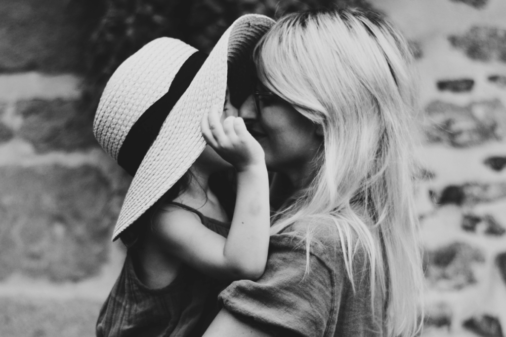 A black and white photo of a woman in a hat lovingly kisses her daughter, capturing a tender moment between mother and child.