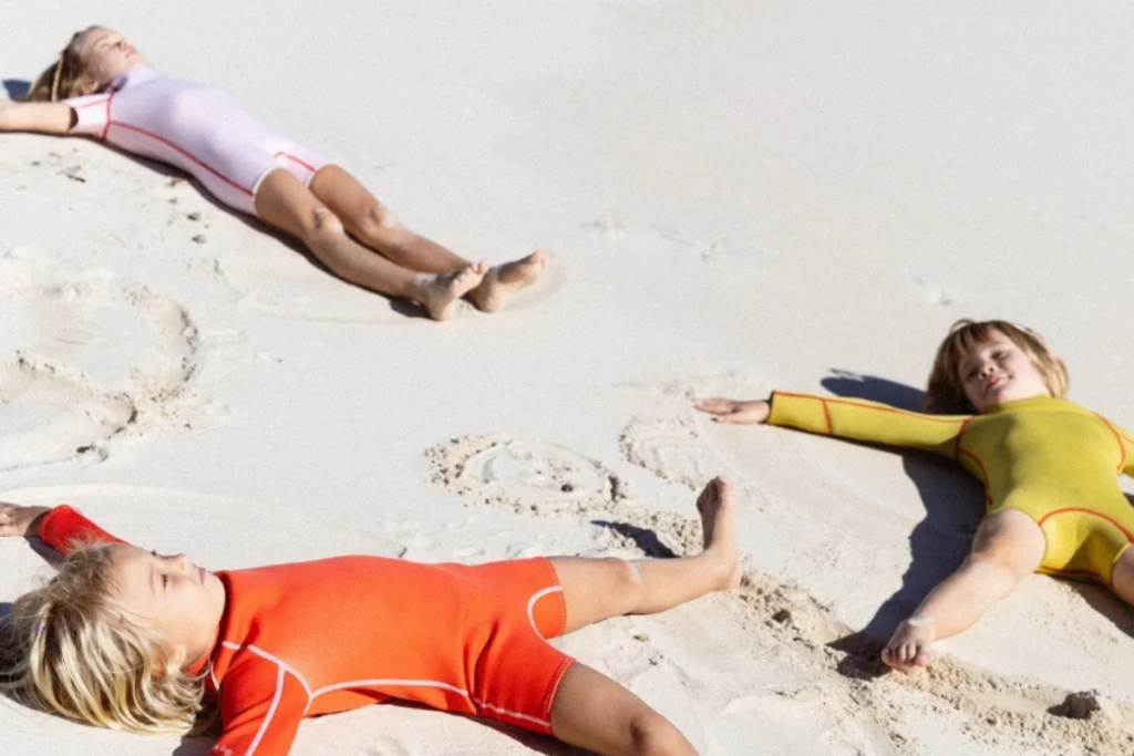 Three children in wetsuits relaxing on the sand, enjoying a sunny day at the beach.