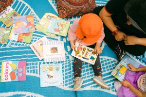 A woman and two children sit on a blanket, engrossed in reading their books outdoors.