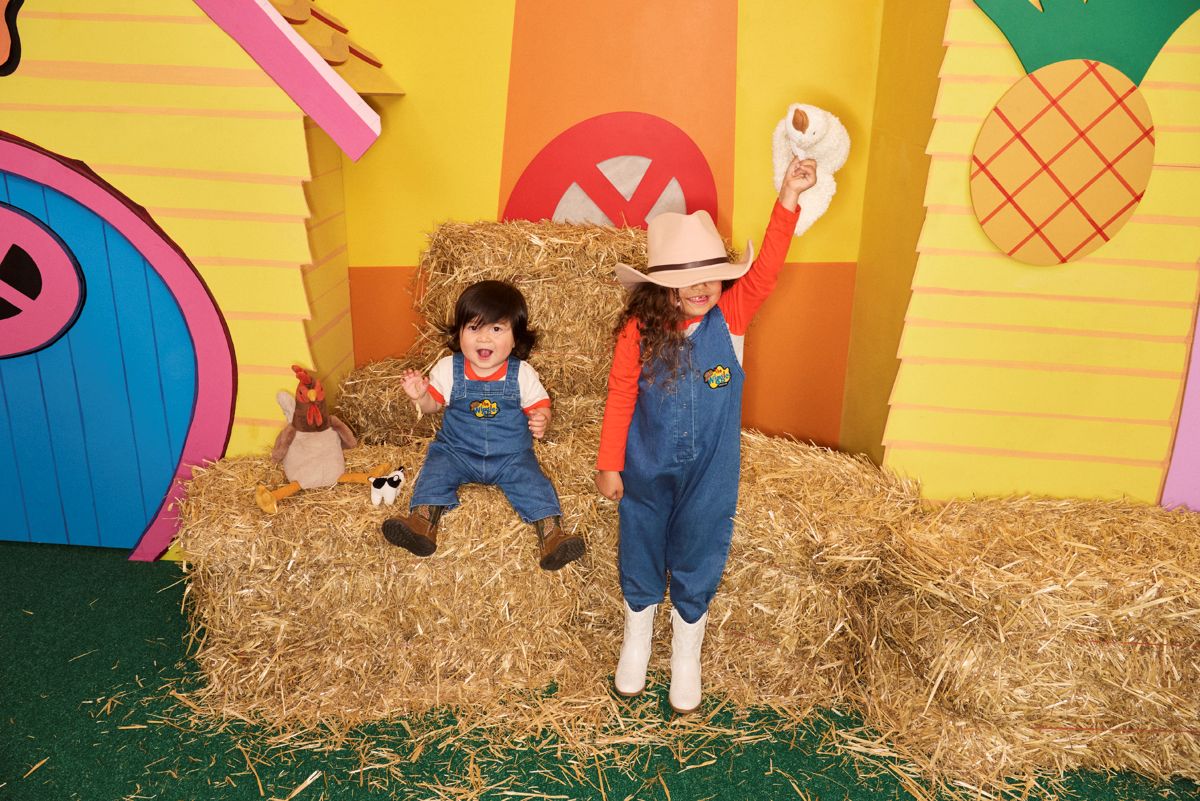  Two children in overalls and cowboy hats smile while posing in front of a rustic barn.