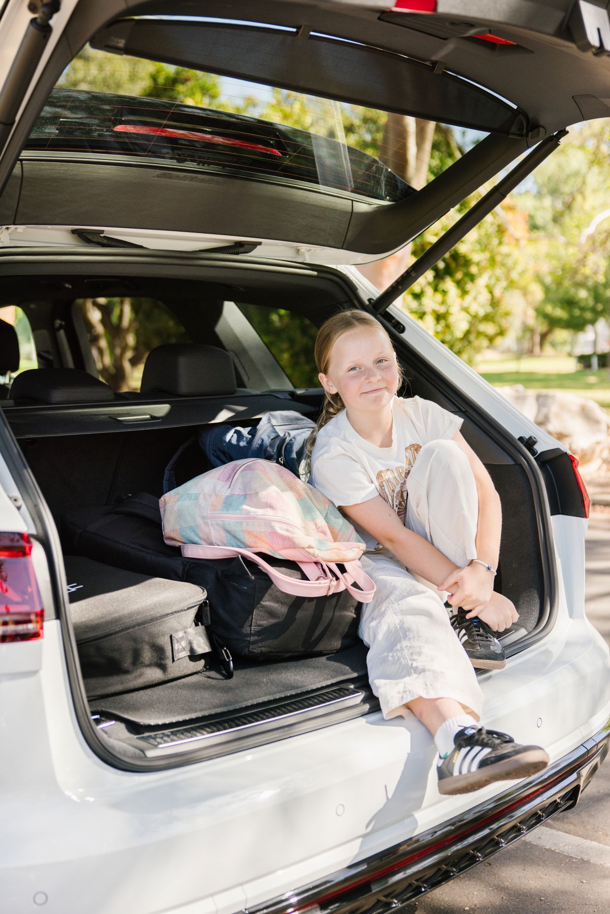 A cheerful young girl is seated in the trunk of a car, surrounded by open space and bright sunlight.