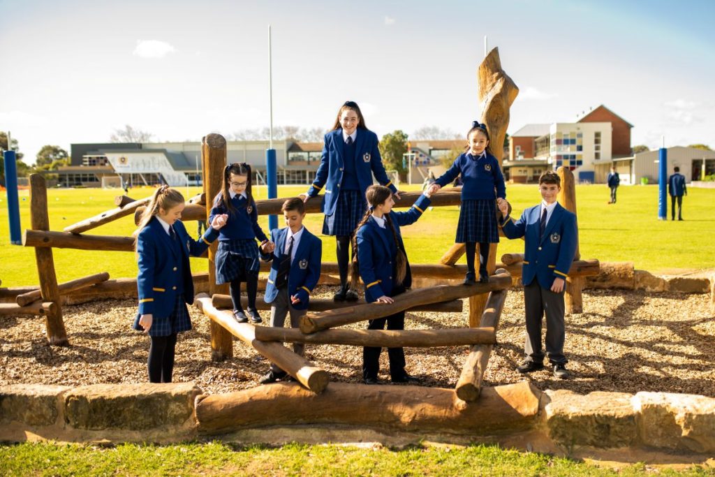 A group of children in school uniforms standing on a wooden structure, smiling.