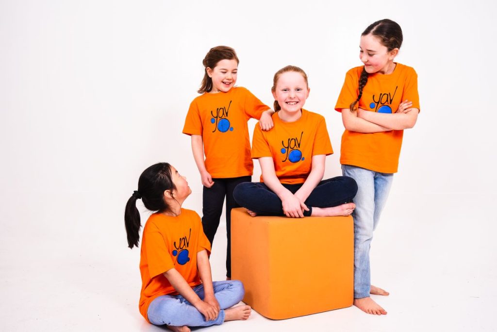 Four children in orange t-shirts sitting on a large cube, smiling and enjoying their time together.