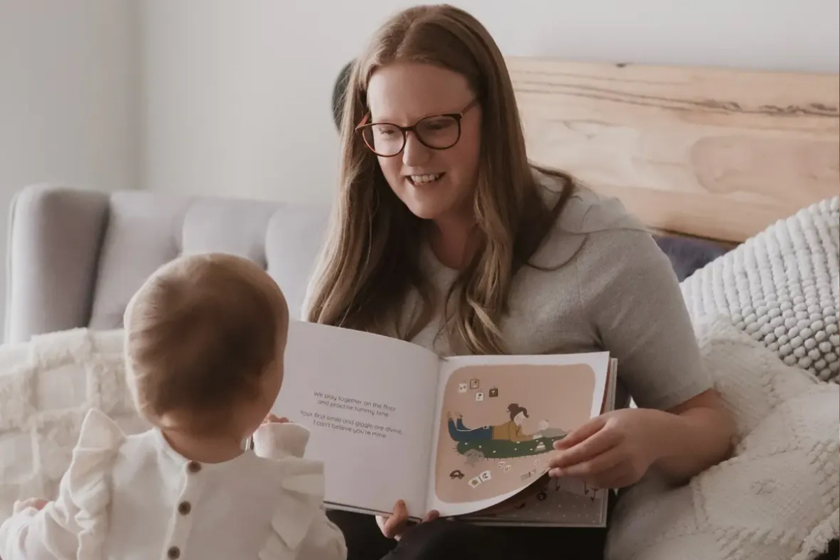 A woman wearing glasses reads a story to a baby nestled comfortably in a bed.