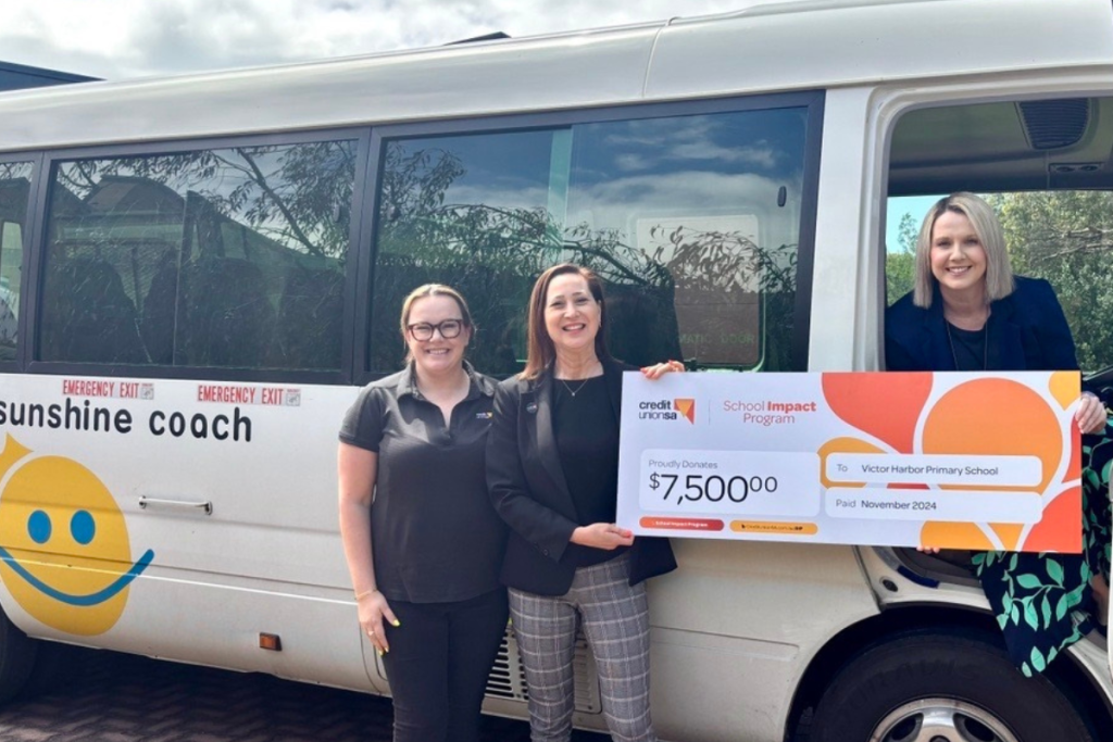 Three women stand beside a bus, each holding a large check, smiling and celebrating their achievement together.