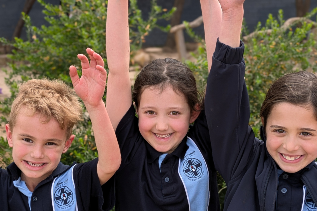 Three children in school uniforms enthusiastically raising their hands in a classroom setting.