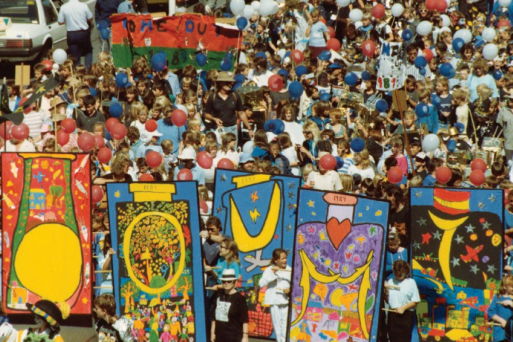 A vibrant crowd gathers, holding colorful banners that create a lively and festive atmosphere.