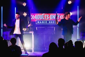 Two men performing on stage with a sign reading "Adults" for a magic show, engaging the audience with their act.