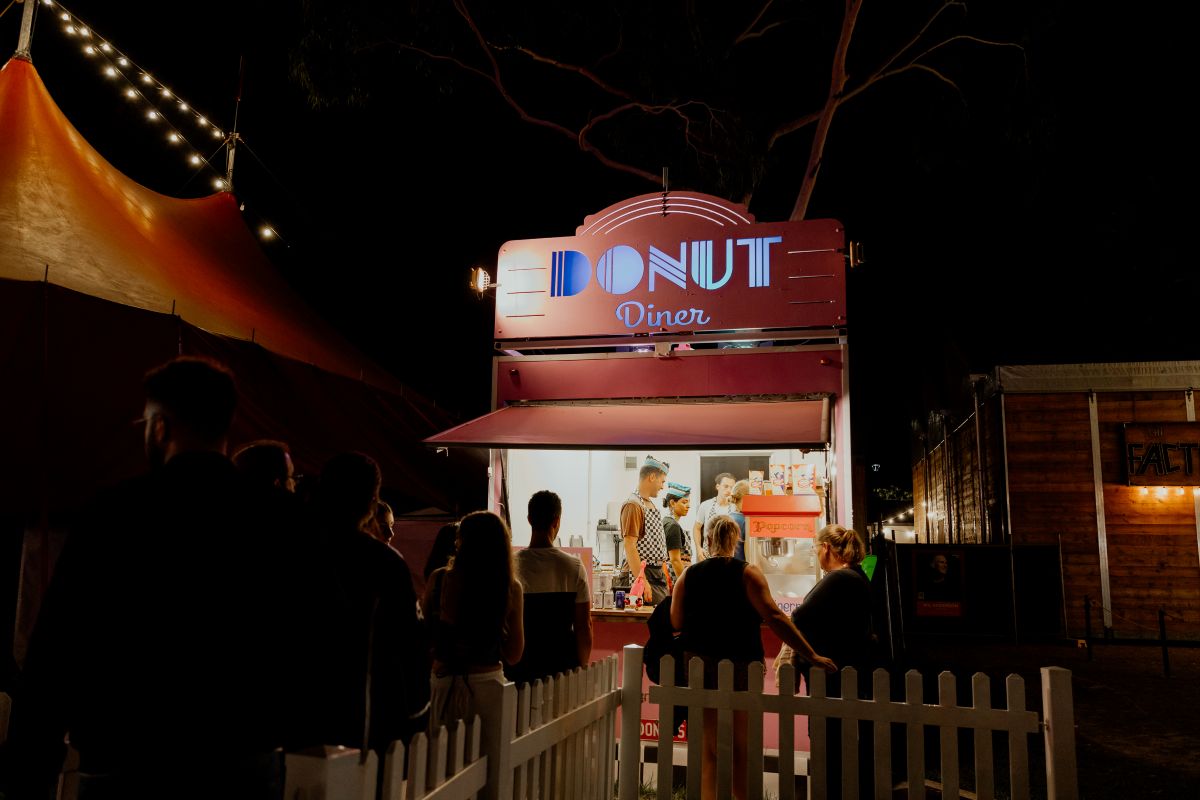 A group of people gathered at a donut stand, illuminated by soft lights, enjoying treats on a lively night.