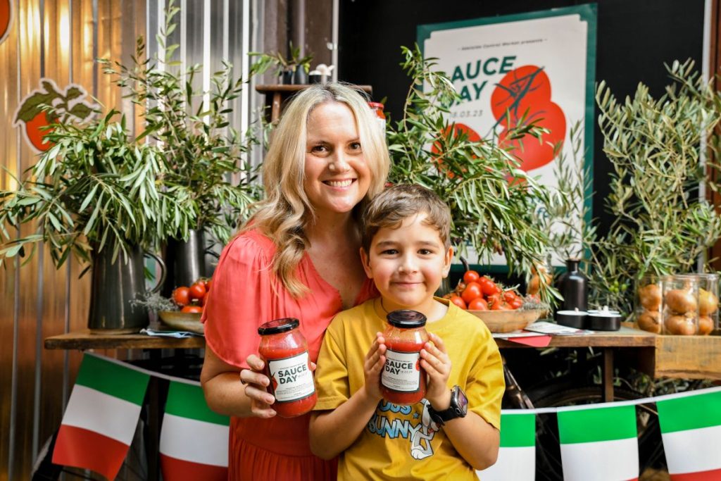 A woman and a boy proudly hold jars of homemade tomato sauce, smiling together in a bright kitchen setting.