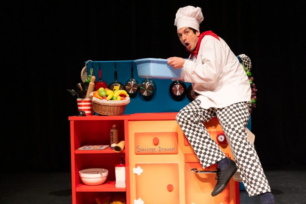 A man in a chef's outfit sits playfully on a toy kitchen, showcasing a whimsical culinary scene.