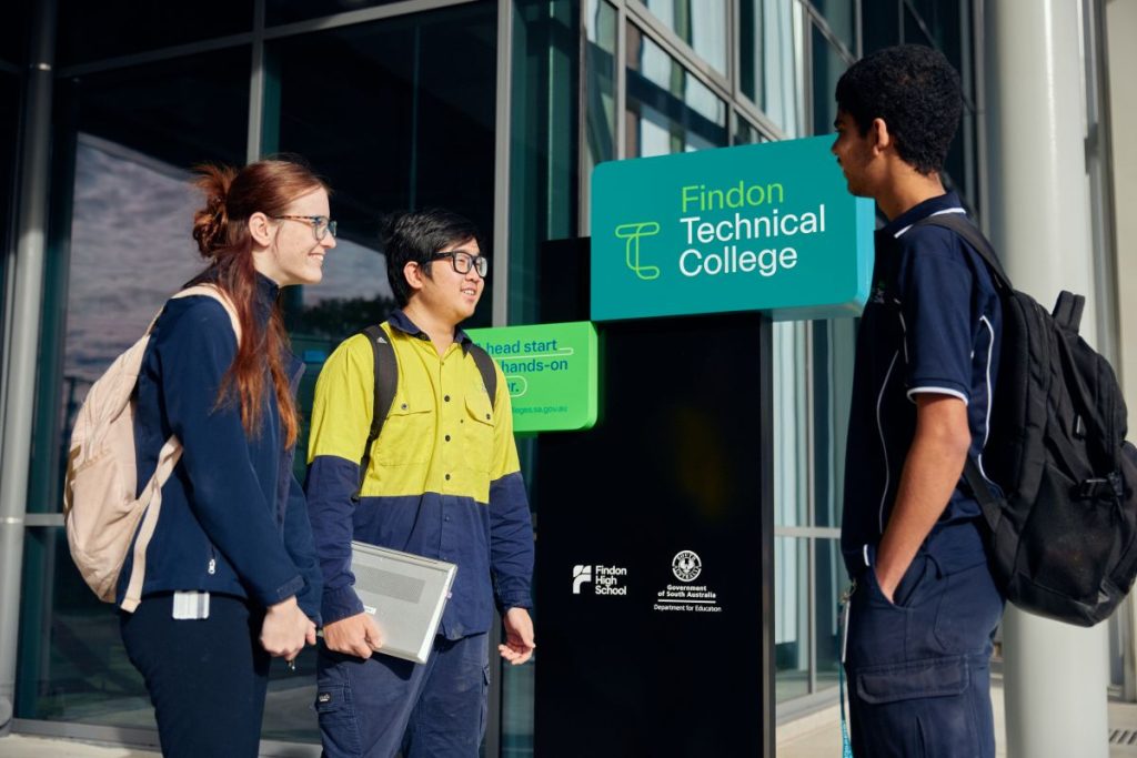 Three students stand outside a building with a sign reading "Technical College," smiling and engaged in conversation.