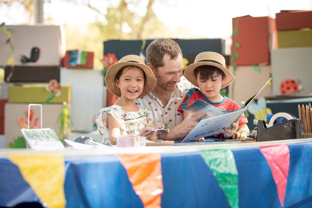 A man and two children are seated at a table, engaged in reading a book together.