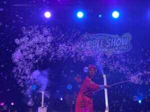 A woman in a flowing red dress joyfully performs, surrounded by shimmering bubbles in a vibrant setting.