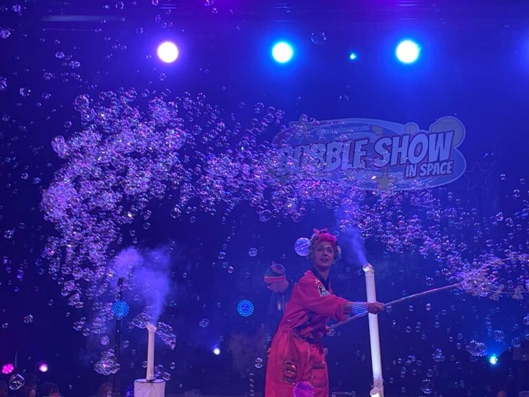A woman in a flowing red dress joyfully performs, surrounded by shimmering bubbles in a vibrant setting.