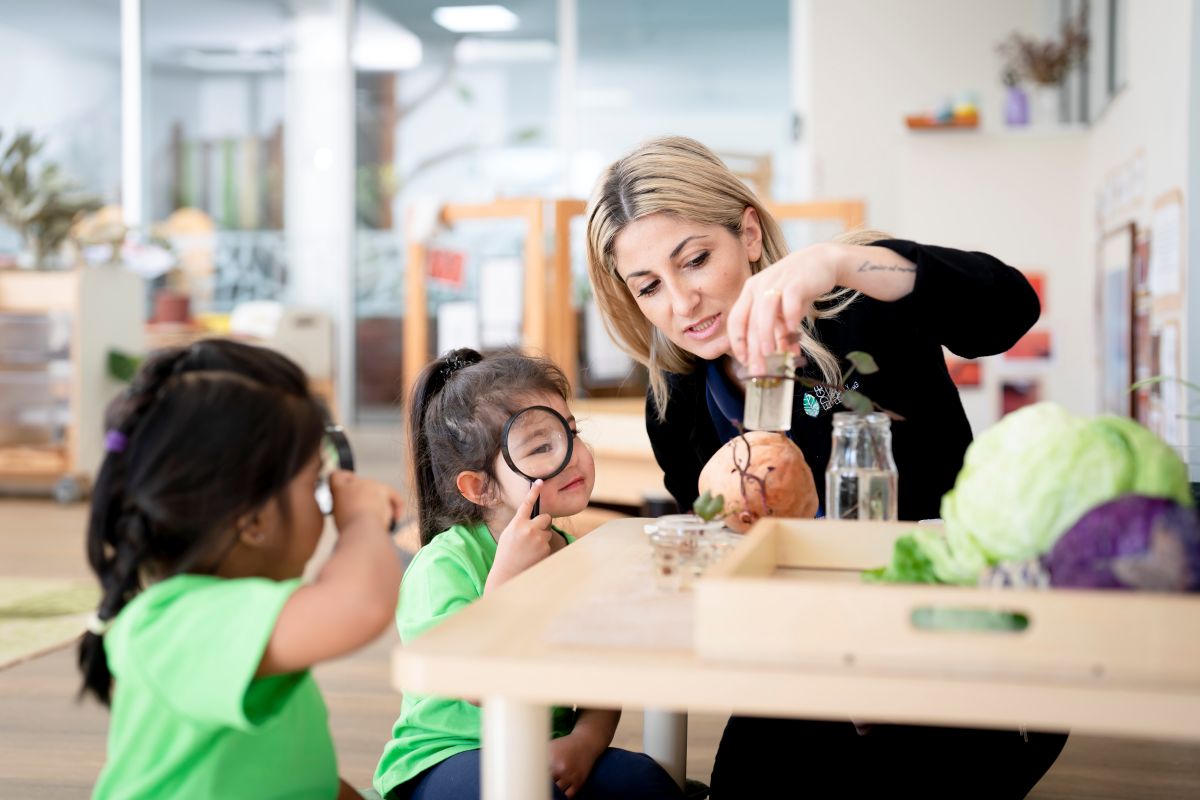 A woman instructs children on vegetable gardening techniques in a sunny outdoor setting.