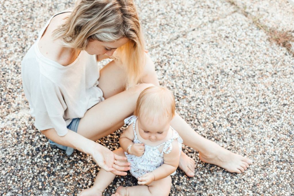 A woman sits on the ground with her baby, both looking content and relaxed in a serene outdoor setting.