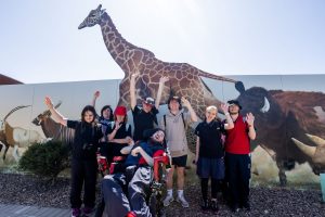 A diverse group of people in wheelchairs gathered together with a giraffe in a sunny outdoor setting.