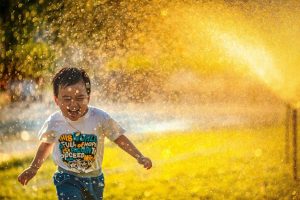 A young boy joyfully runs through a sprinkler, water spraying around him on a sunny day.