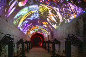 A vibrant tunnel illuminated with colorful lights, featuring a large projection screen at the end.