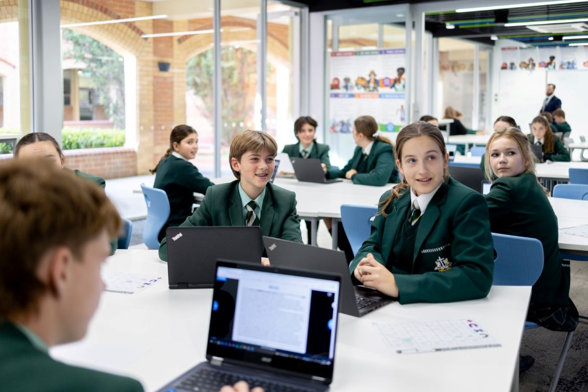 A group of students in school uniforms working on laptops at tables in a classroom setting.