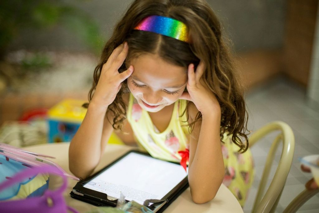 A young girl sits at a table, focused on her tablet, with a curious expression on her face.