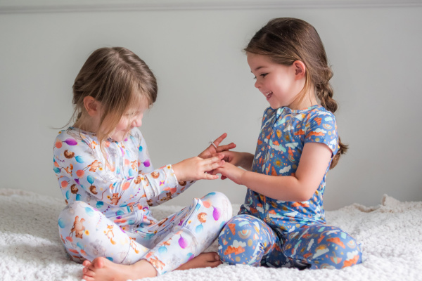 Two young girls sitting on a bed, smiling and resting their hands on each other's arms.