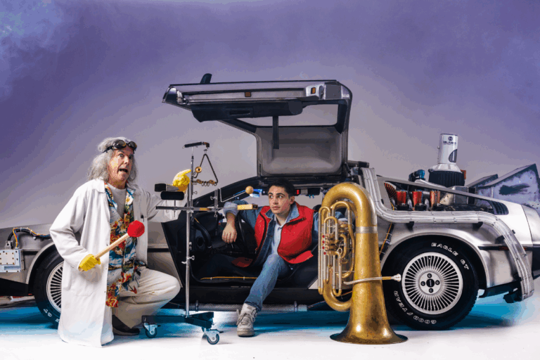 Two people in colorful costumes pose beside a car, one holding a trumpet, smiling for the camera.