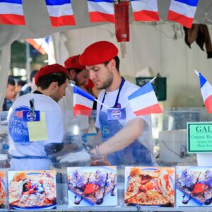 A man in a red hat and shirt sells French food at a market stall.