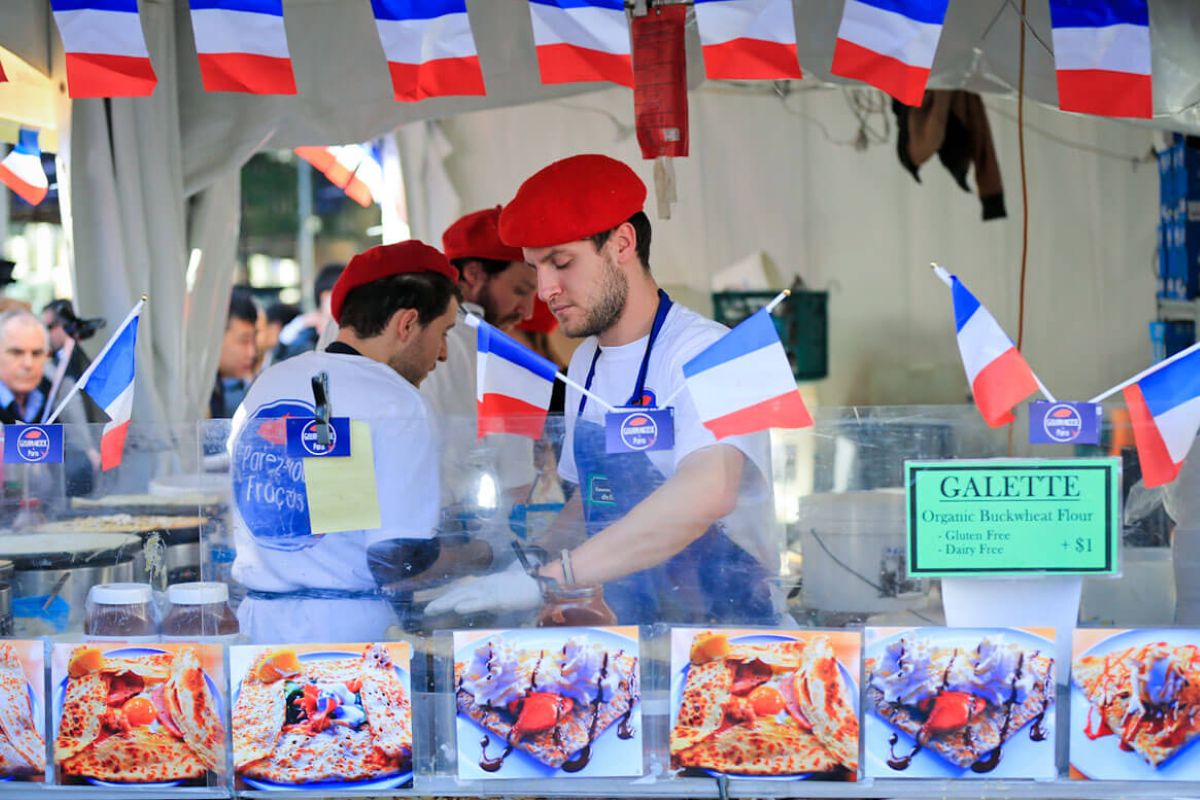 A man in a red hat and shirt sells French food at a market stall.