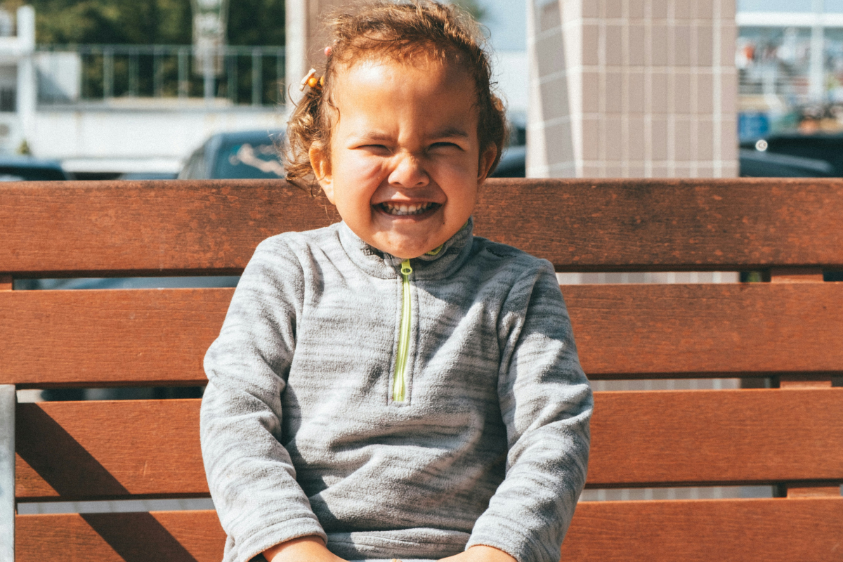 A little girl sits on a bench, laughing joyfully with her hands on her knees.
