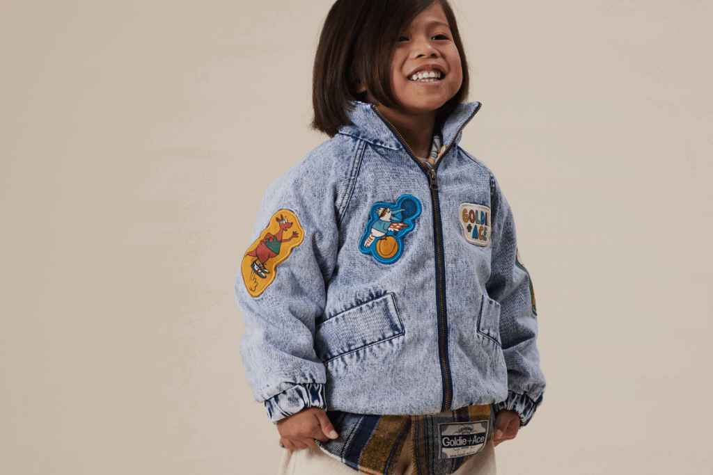 A young girl in a denim jacket adorned with colorful patches, smiling at the camera.