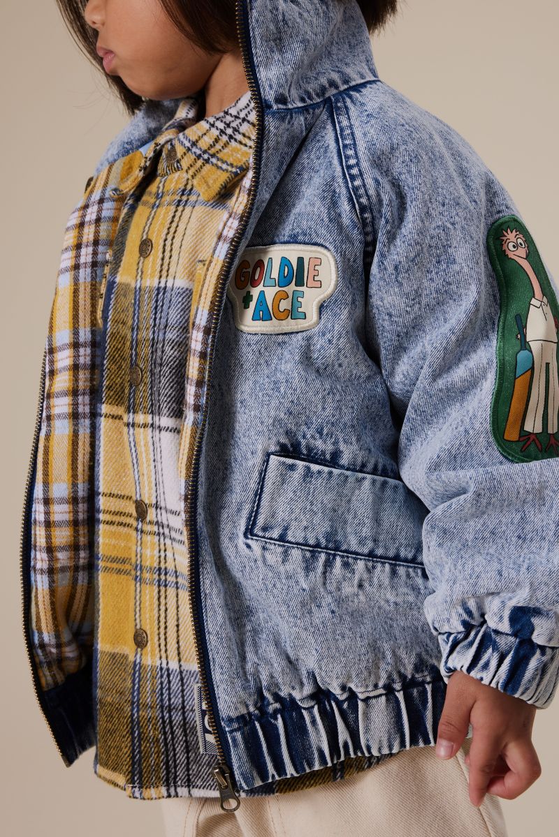 A young child in a denim jacket over a plaid shirt, smiling and standing outdoors. 