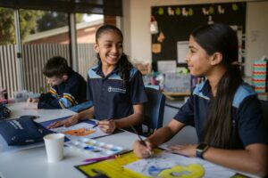 Two girls in school uniforms sit at a table, surrounded by various art supplies, engaged in creative activities.