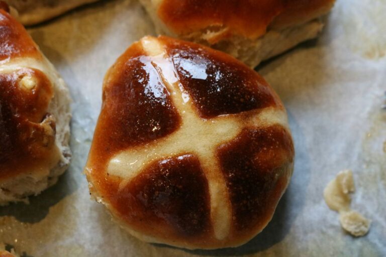 Freshly baked hot cross buns with a golden-brown crust and a white cross on top, arranged on a wooden surface.
