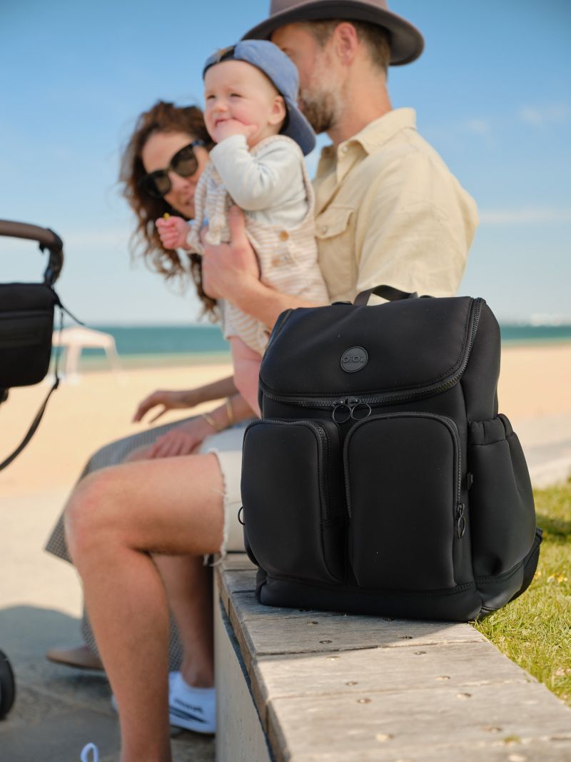 A man and woman sit on a bench beside a stroller with a baby, enjoying a sunny day in the park.